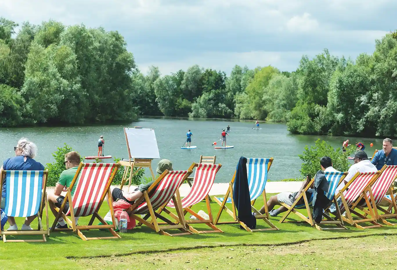 People sitting in deckchairs by the lake, watching paddleboarders