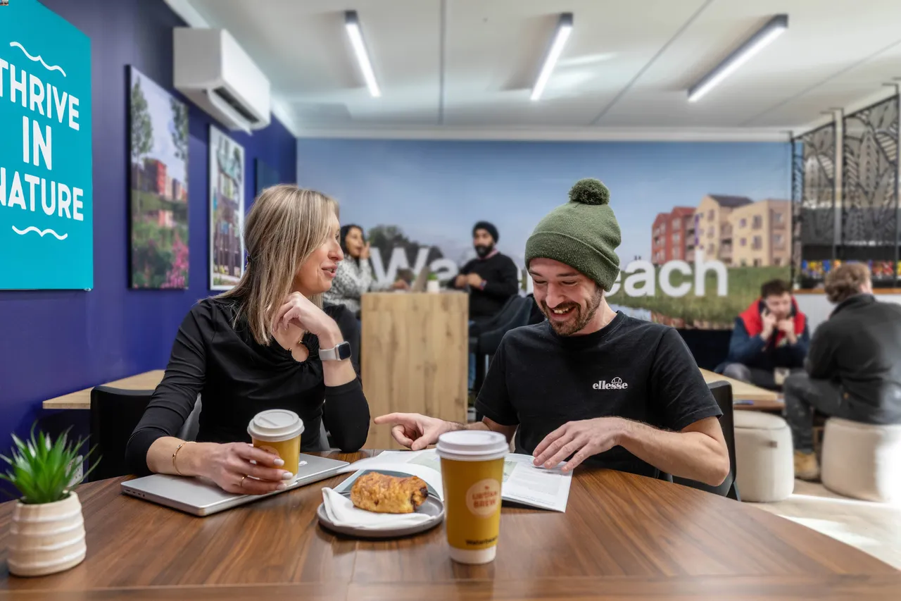 Two people sit at a wooden table, smiling and talking over coffee and pastries. A wall sign reads "Thrive in Nature," and others converse in the background.