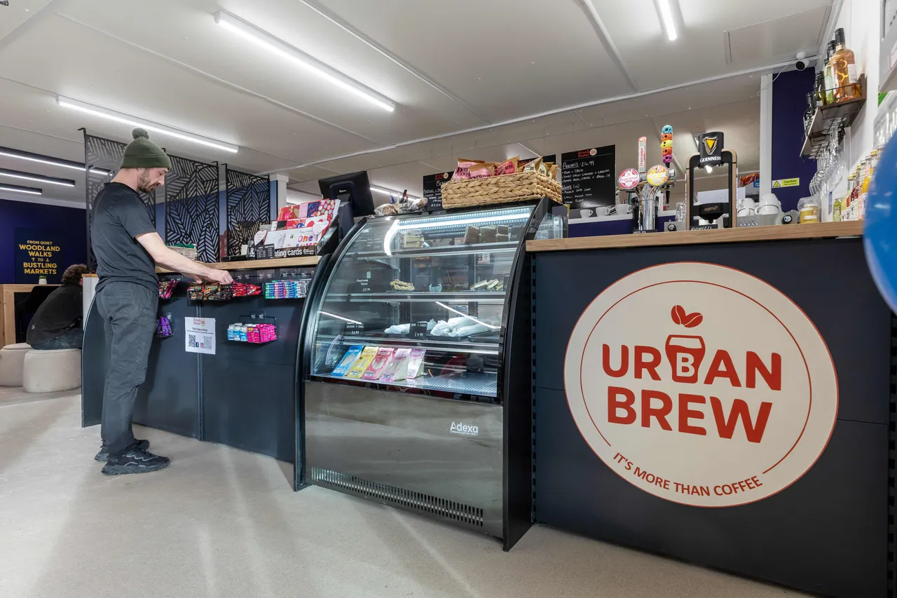 A man in a beanie looks at snacks on the counter of a cozy café. A sign reads "Urban Brew" on a display case filled with treats and drinks.