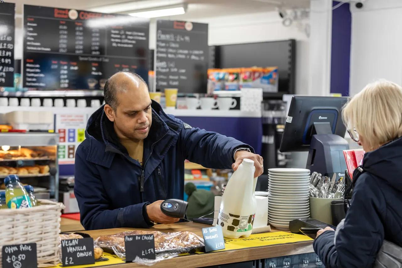 A man scans a milk bottle at a café counter while a woman stands on the other side holding a phone. The café displays menus, pastries, and utensils.