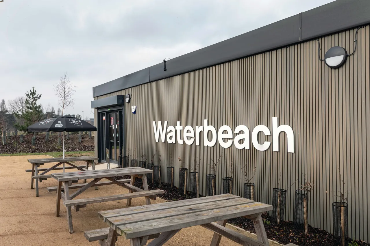 Outdoor seating area with wooden picnic tables and black umbrellas near a building marked "Waterbeach." Overcast sky gives a calm, inviting tone.