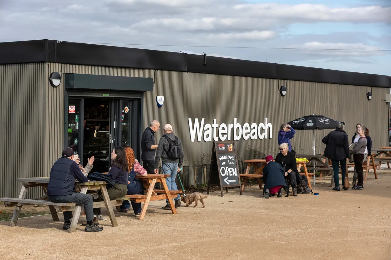 A group of people sits outside the Waterbeach Cafe, enjoying coffee and sunshine on a pleasant day.