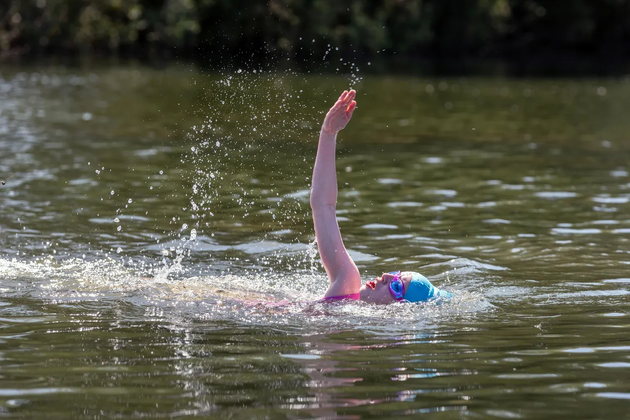 A close-up action shot of a person swimming backstroke in a lake. Their arm is extended straight up toward the sky, creating a spray of water droplets against the sunlight. They are wearing pink swimwear, blue goggles, and a blue swim cap.