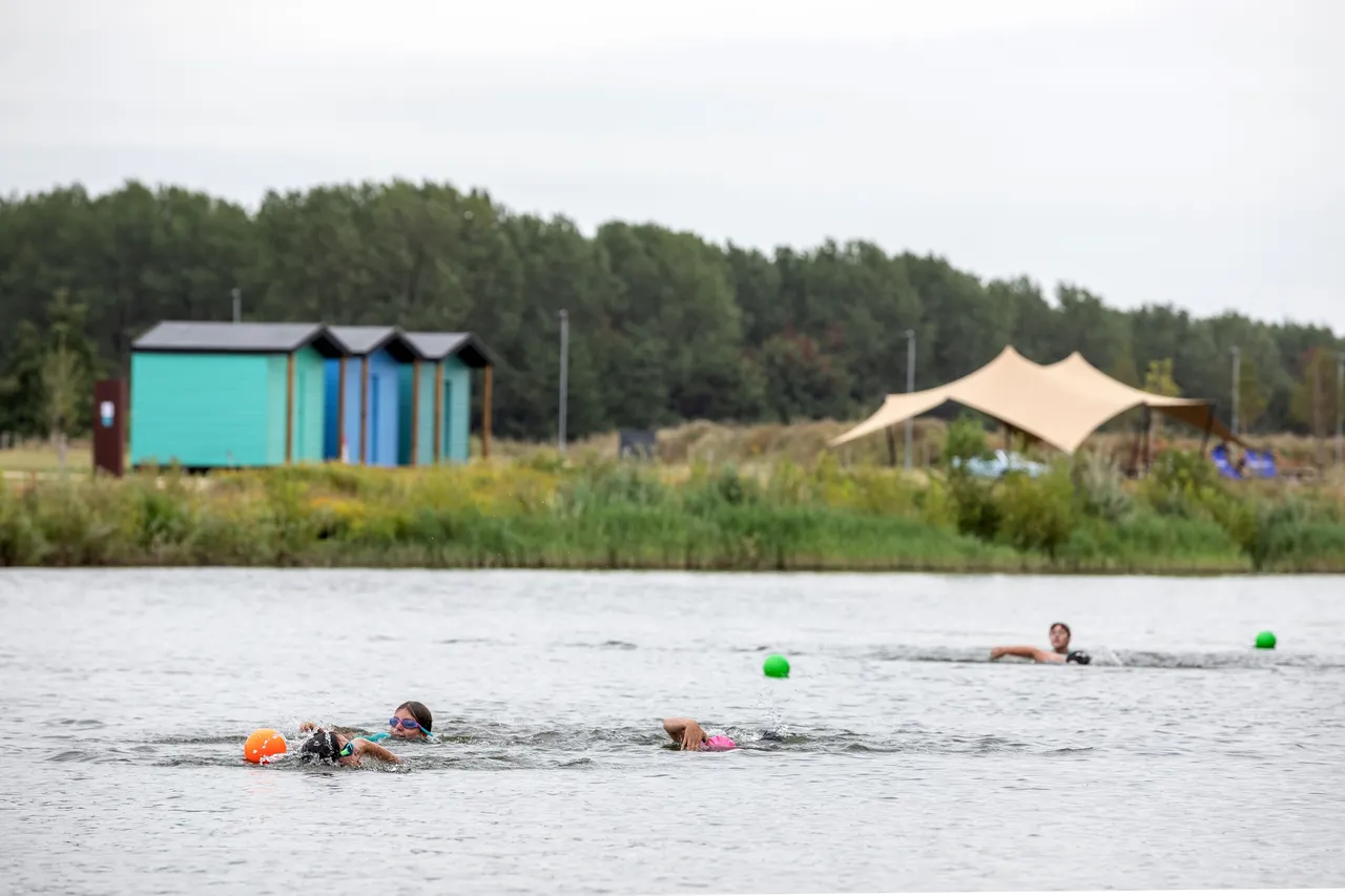 A group of people swimming in a large lake under an overcast sky. Brightly colored buoys (orange and green) float on the surface to mark the swimming course. In the background, there are small turquoise and blue changing huts and a large tan sunshade structure near the treeline.