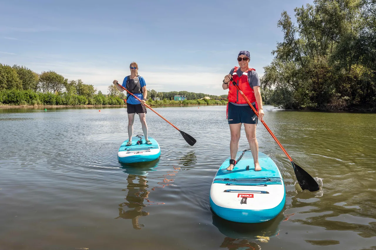 Two people standing on blue and white paddleboards in the middle of a lake on a bright, sunny day. Both are holding long paddles and wearing life jackets and sunglasses. The water is calm, reflecting the clear blue sky and the lush green trees along the shore.