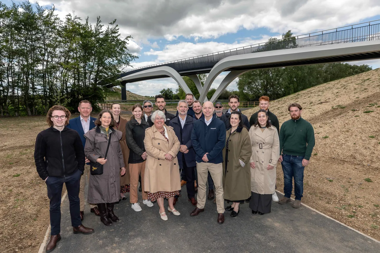 A wide group portrait of approximately 18 people standing on a paved path directly beneath the A10 bridge. The bridge features a striking modern design with a curved, white Y-shaped support pillar and a dark metal walkway above. The group is dressed in smart-casual attire, including coats and jackets, posing against a backdrop of green trees and a grassy embankment under a cloudy sky.