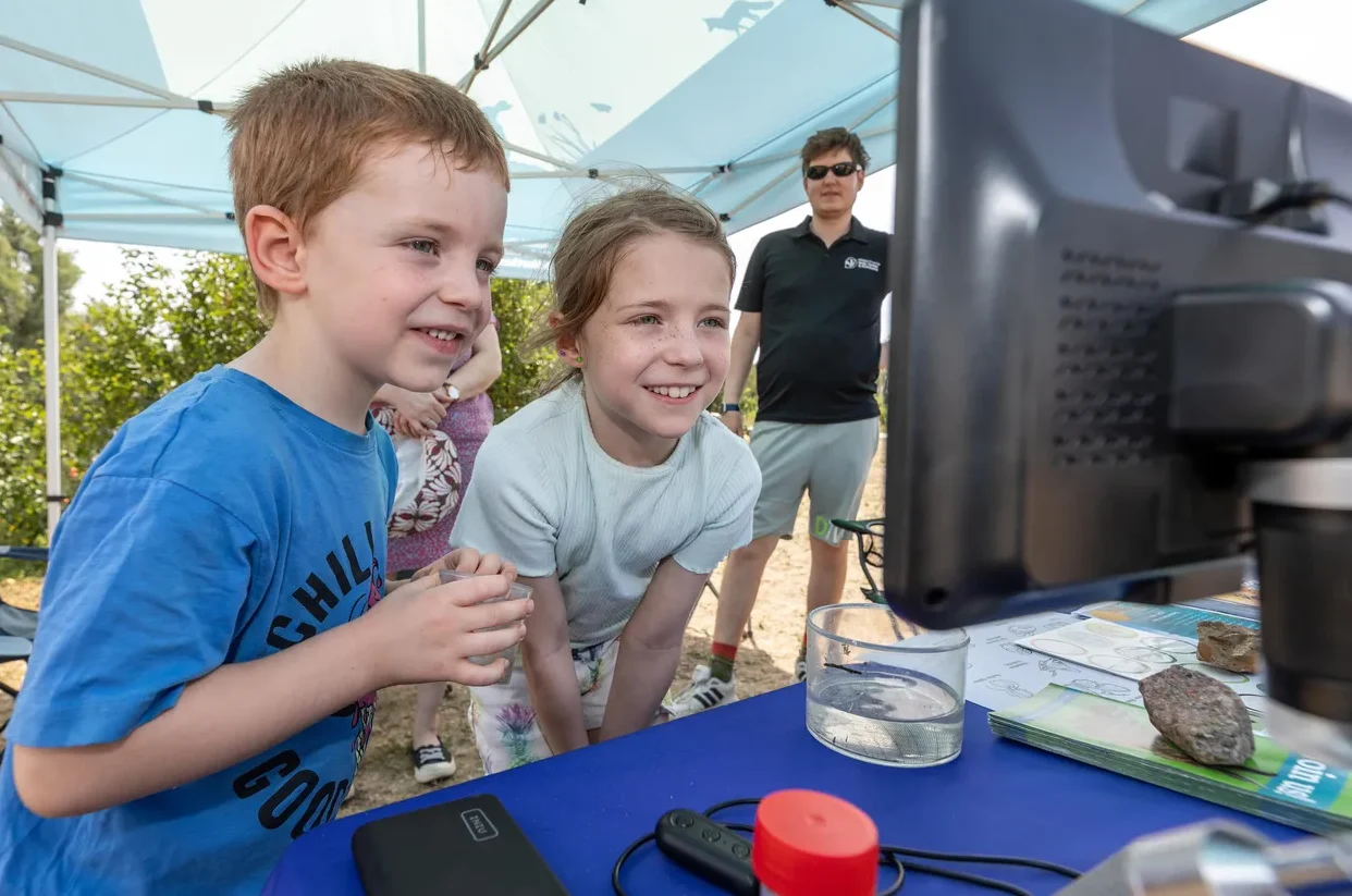 Two young children smiling bright as they look closely at a digital display screen showing magnified pond organisms under a canopy tent, with water samples and identification sheets on the table in front of them.