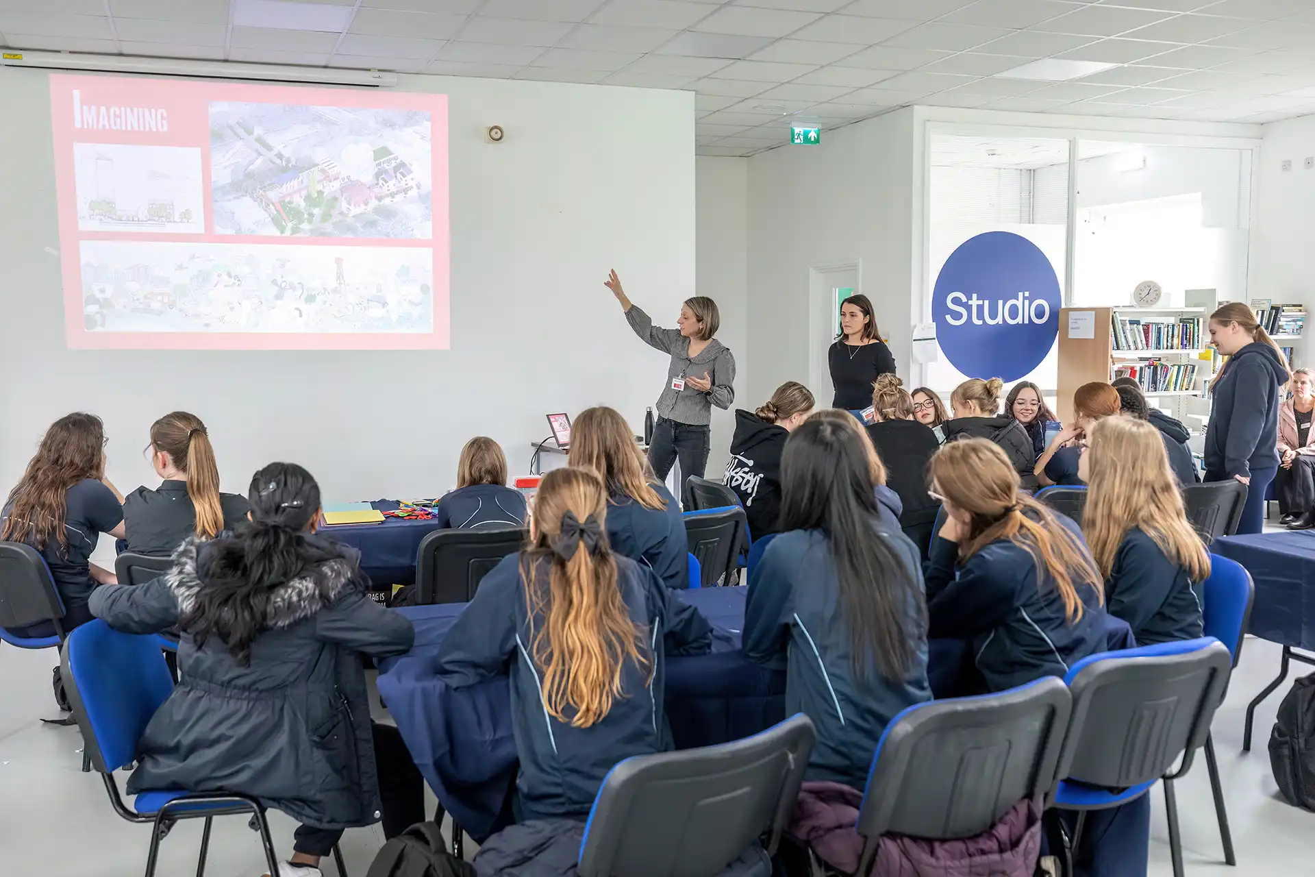 School students in navy uniforms attend a workshop, listening to a presenter beside a projected slide showing urban planning illustrations